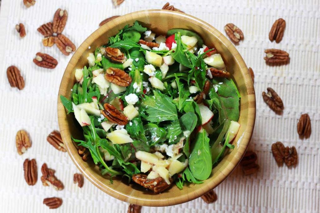 A wooden bowl of mixed arugula salad topped with candied pecans, diced apples, and goat cheese crumbles.