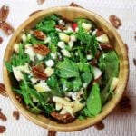 A wooden bowl of mixed arugula salad topped with candied pecans, diced apples, and goat cheese crumbles.