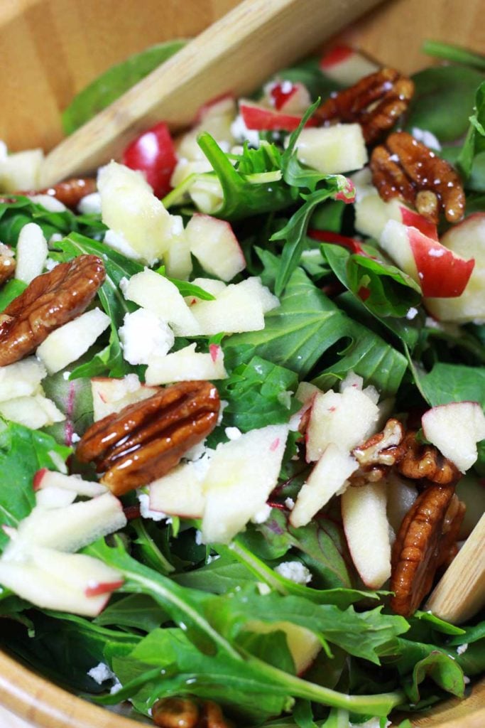 A close up of arugula salad topped with candied pecans, diced apples, and goat cheese crumbles.