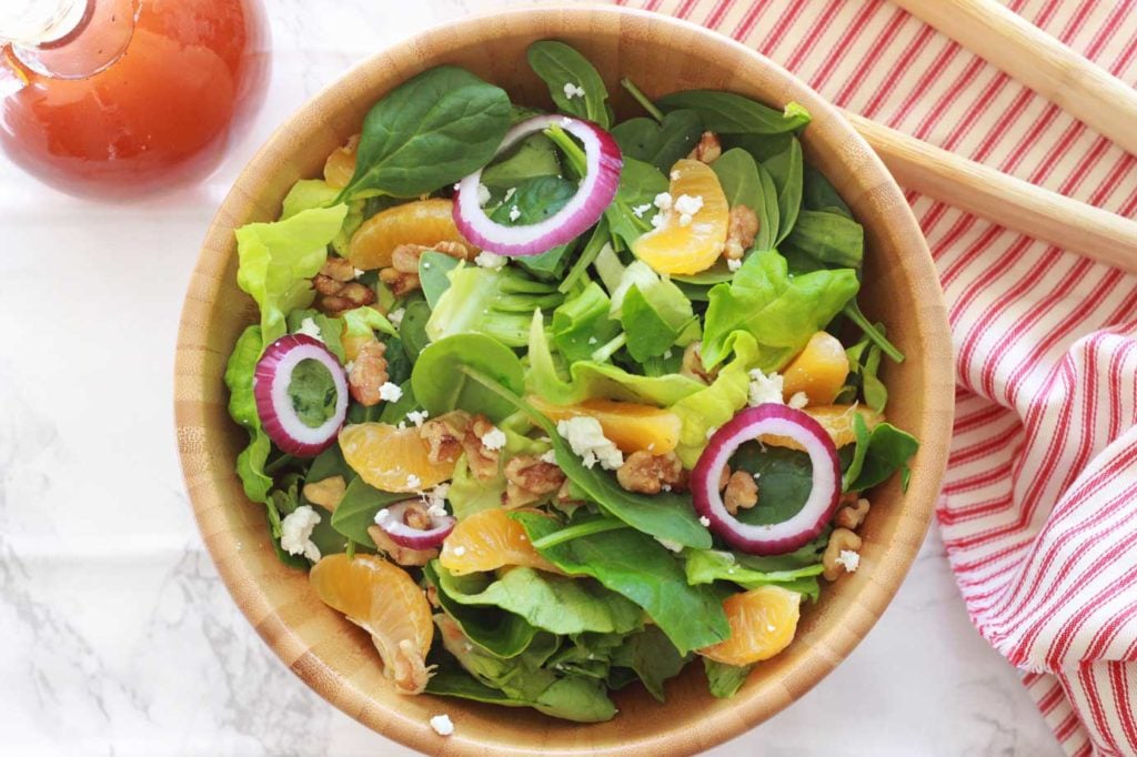 A wooden bowl of green salad topped with red onions, walnuts, and goat cheese crumbles.