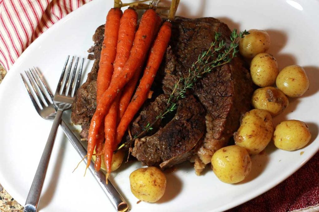A plate of a pot roast with whole carrots, potatoes, and thyme sprigs with a fork.