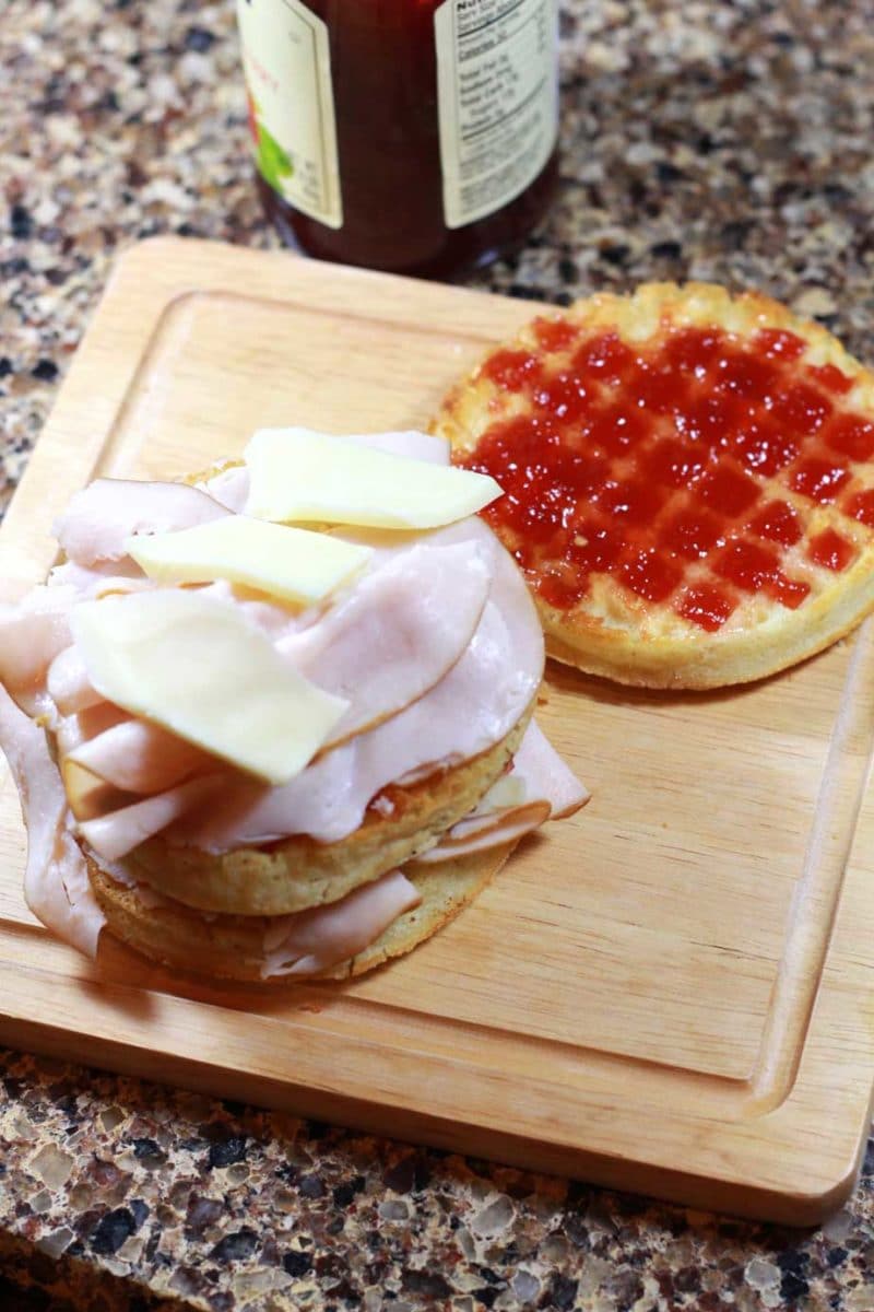 Wooden cutting board with a layered waffle containing cheese, turkey and raspberry jam being used to make a Monte Cristo.