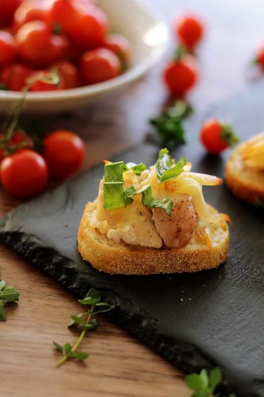 Black slate plate containing a Chicken Baguettes topped with Gruyère cheese and basil, heirloom tomatoes in a white bowl.