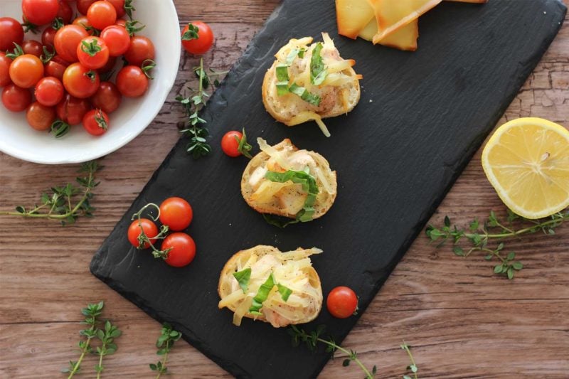 Three Garlic Mayo Chicken Baguettes with Gruyère cheese on a black slate plate, sitting on a wooden table with thyme sprigs, lemons and heirloom tomatoes.