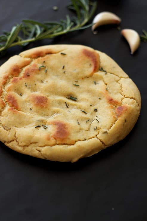 A Garlic Flatbread round topped with fresh rosemary sitting on a black table, rosemary and garlic in background.