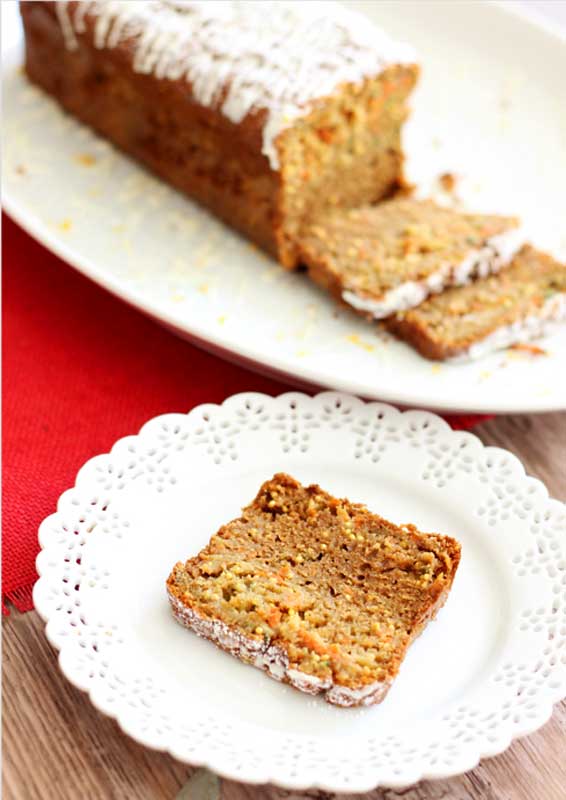 Zucchini, Carrot and Apple Olive Oil Cake topped with cream cheese frosting sitting on a white lace plate on wooden table, loaf of cake in background.