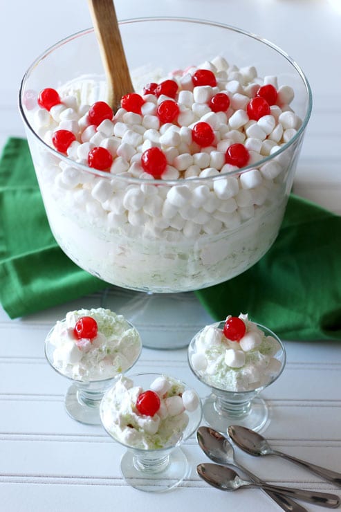 Trifle bowl containing a Pineapple Pistachio Marshmallow Dessert topped with cherries, 3 small parfait glasses will dessert, 3 spoons on table.