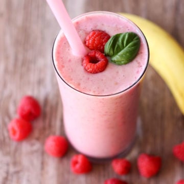 Glass containing a berry smoothie topped with 2 raspberries and a mint spring, berries and banana on table.