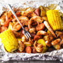 Tin foil containing sauteed shrimp and corn on the cob, shrimp fork serving.