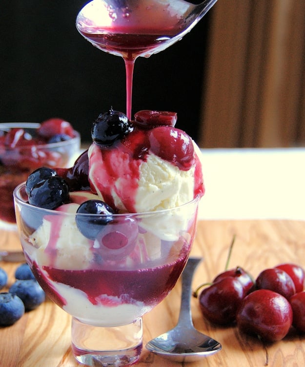 Cheesecake ice cream in a parfait glass, cherry and blueberry sauce being poured onto ice cream.