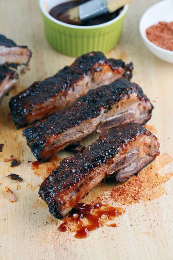 Wooden table containing 3 BBQ ribs, green ramekin with BBQ sauce and brush.