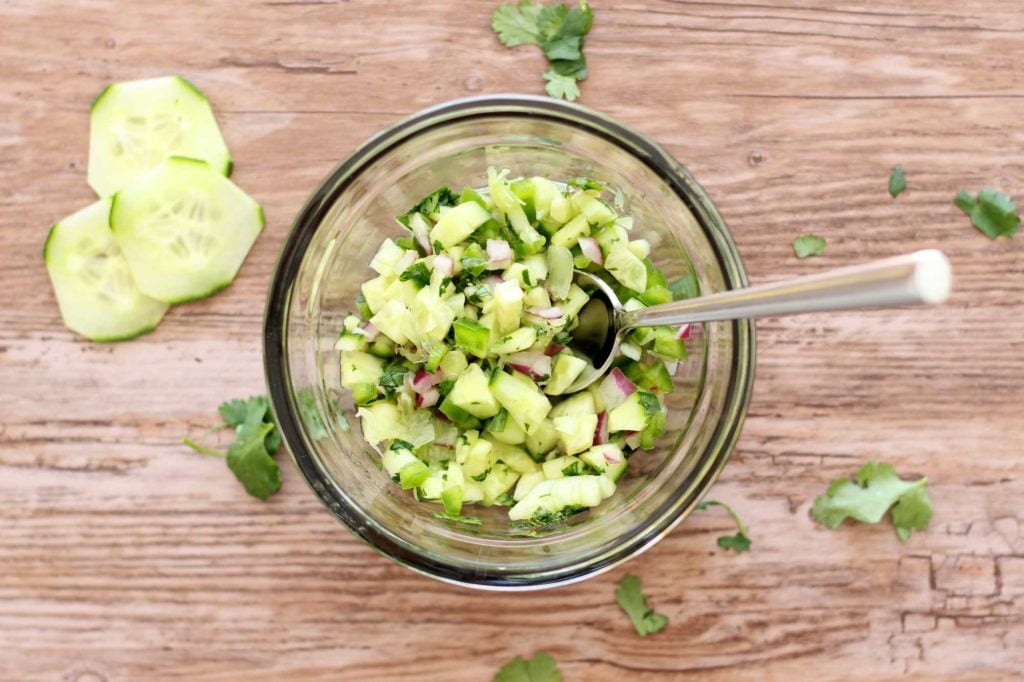 Clear bowl containing cucumber lime salsa sitting on a brown tabletop, cucumbers and cilantro on table.