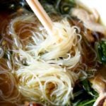 White bowl of Asian Chicken Noodle Soup with Mei Fun Noodles twirled on chopsticks, sitting on a brown table mat.