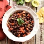 White bowl containing a black bean dip containing tomatoes and corn, lime and tortilla chips on wooden table.