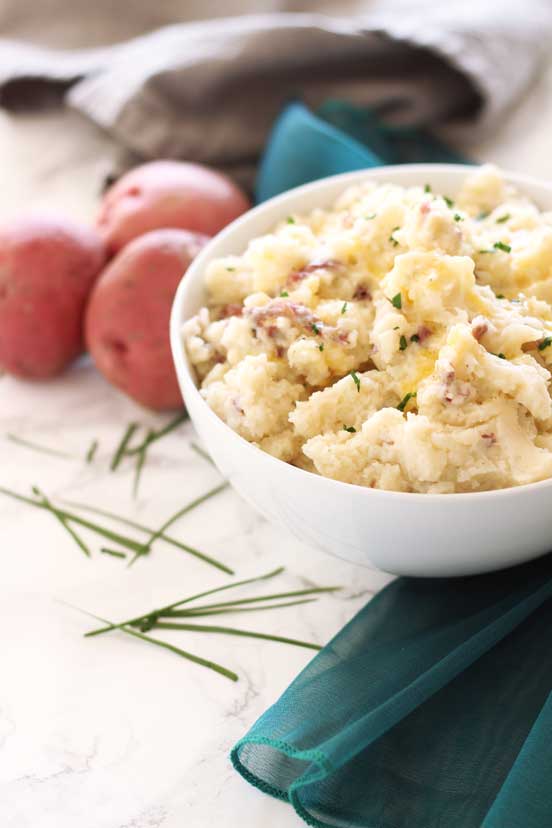 White bowl containing homemade mashed potatoes topped with chives, sitting on a white table with a teal napkin, red potatoes in background.