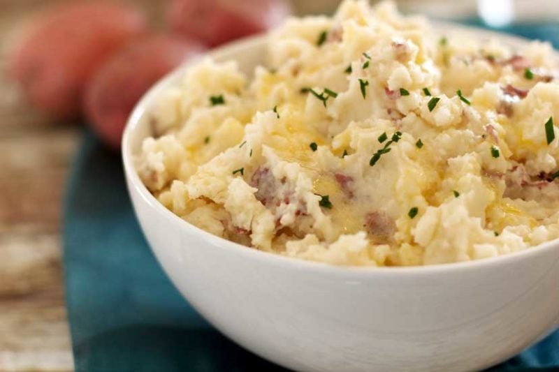 White bowl containing mashed potatoes topped with chives sitting on a wooden table, teal napkin and red potatoes in background.