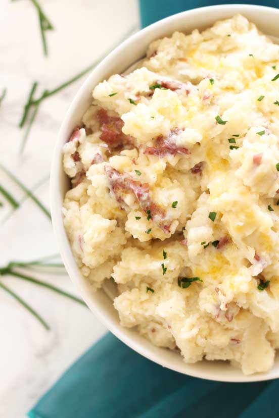 White bowl containing homemade mashed potatoes sitting on a white table with a teal napkin, chives scattered on table.