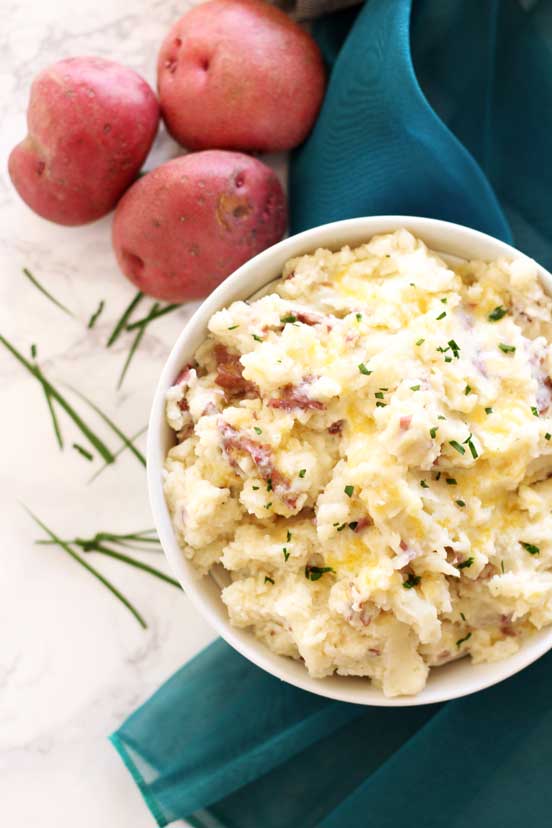 Bowl of homemade mashed potatoes in a white bowl sitting on a marble table; teal napkin, chives and 3 red potatoes in background.