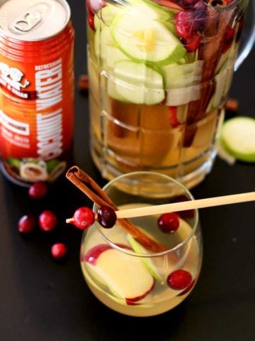 Short glass containing Cinnamon Apple White Sangria topped with a cinnamon stick and cranberries, can of Amy and Brian's coconut water and pitcher on black table.