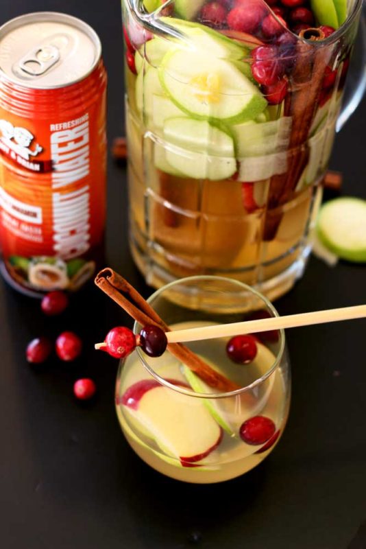 Short glass containing Cinnamon Apple White Sangria topped with a cinnamon stick and cranberries, can of Amy and Brian's coconut water and pitcher on black table.