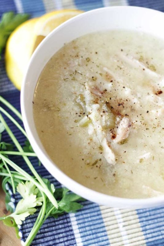 A white bowl containing Greek Lemon and Chicken Soup (Avgolemono Soup) sitting on a blue tablecloth, parsley and lemons on table.