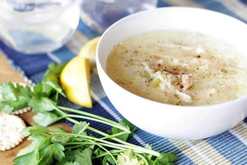 A white bowl containing Greek Lemon and Chicken Soup (Avgolemono Soup) sitting on a blue tablecloth, crackers, parsley and lemons on table.