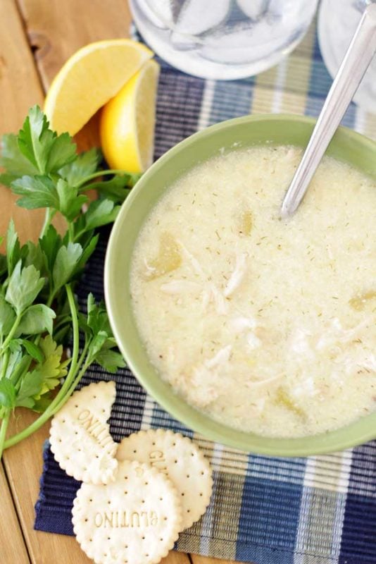 A Green bowl containing Greek Lemon and Chicken Soup (Avgolemono Soup) sitting on a wooden table, cracker, parsley and lemon on table.