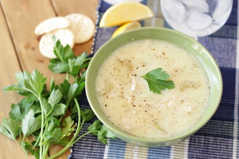 A green bowl containing Greek Lemon and Chicken Soup (Avgolemono Soup) sitting on a wooden table, crackers, parsley, lemon and glass of water on table.