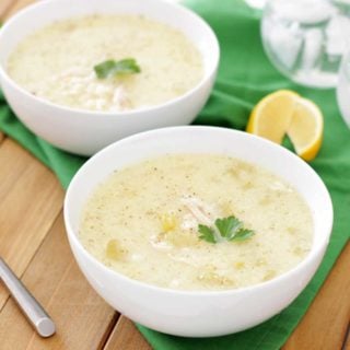 2 white bowls filled with Greek Lemon and Chicken Soup (Avgolemono Soup) sitting on a wooden table, 2 crossed spoons and green napkin on table.