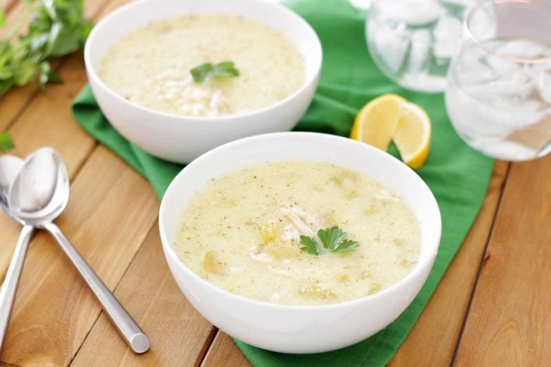 2 white bowls filled with Greek Lemon and Chicken Soup (Avgolemono Soup) sitting on a wooden table, 2 crossed spoons and green napkin on table.