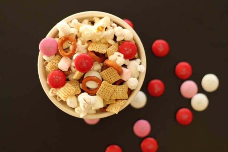 Bowl sitting on a black table containing popcorn mix filled with M&Ms, popcorn, chex and pretzels.