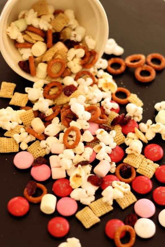 Black table with a spilled bowl of popcorn snack mix containing popcorn, chex cereal, pretzels, dried cranberries and M&M.s