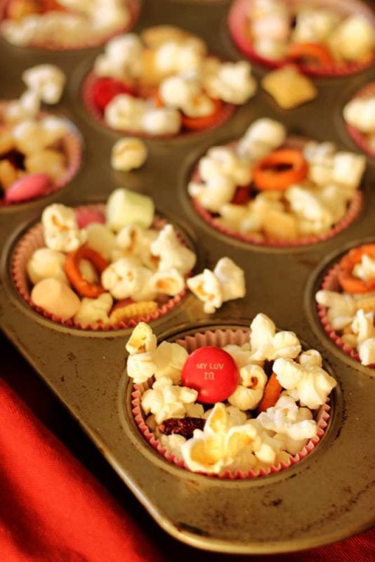 Muffin tin sitting on a red napkin filled with muffin cups of popcorn trail mix containing popcorn, pretzels and M&Ms.