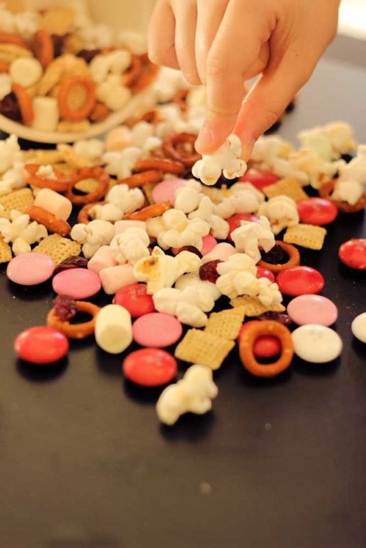 Bowl of popcorn trail mix spilled on table with a child's hand picking up a piece of popcorn.