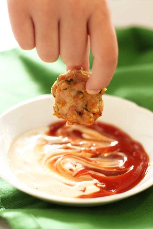 Hand dipping a Zucchini Chicken Bite into a white dish of ranch and ketchup, dish sitting on a green napkin.