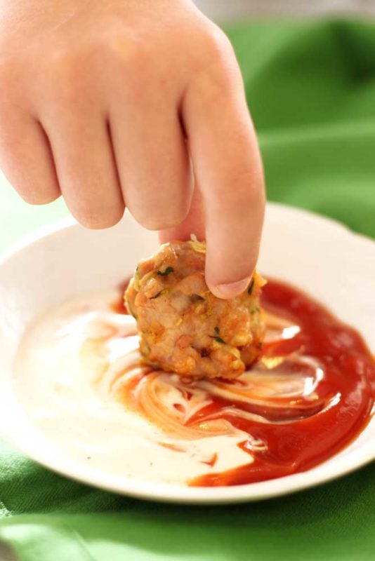 A hand that has dipped a Zucchini Chicken Bite into a white dish of ranch and ketchup, dish sitting on a green napkin.