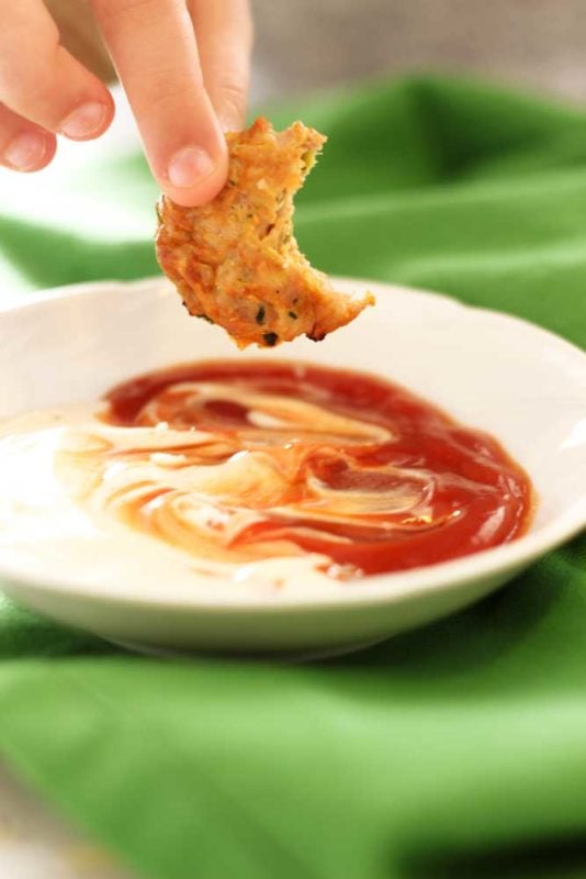 A half eaten Zucchini Chicken Bite into a white dish of ranch and ketchup, dish sitting on a green napkin.