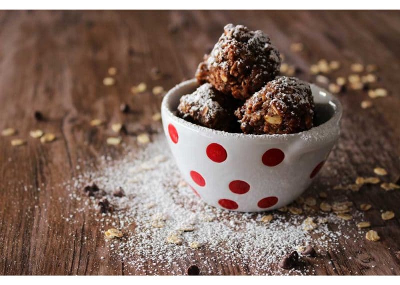 White bowl sitting on a brown table with No-Bake Quinoa Protein bites, oats and powdered sugar on table.