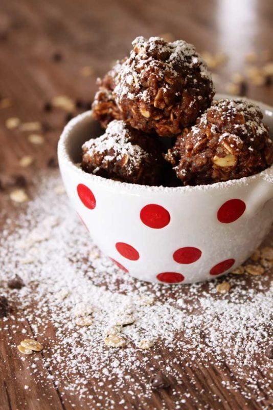 White bowl with red polka dots containing 3 Quinoa Protein Bites sitting on a brown table, sprinkled powdered sugar.