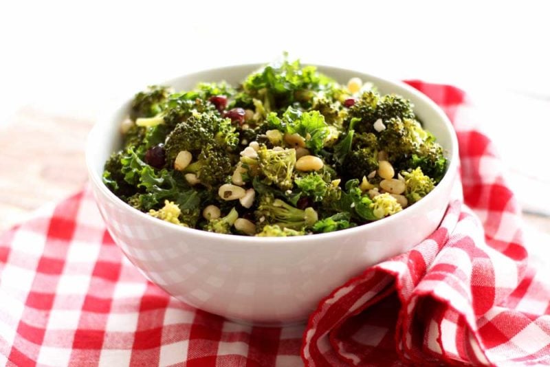 White bowl containing broccoli and kale salad with pine nuts and feta cheese on tip, sitting on a red and white gingham napkin.