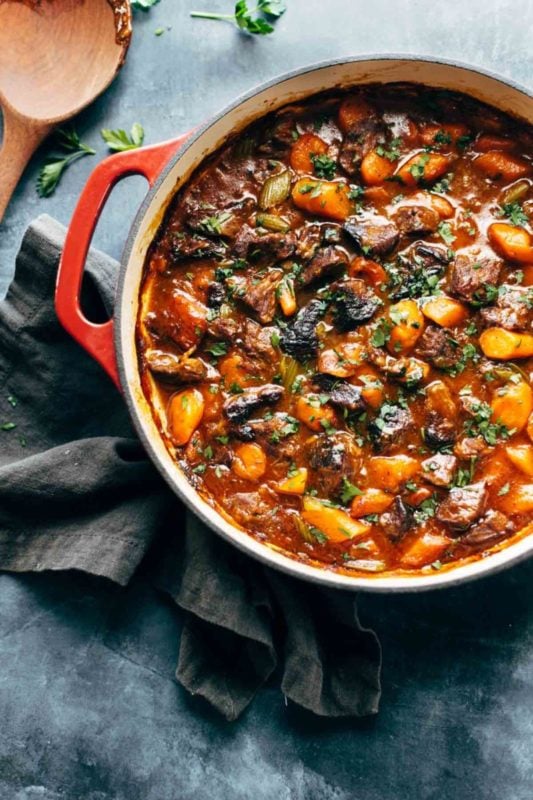 Stew pot containing beef stew with carrots and parsley, sitting on a gray table with a wooden spoon.