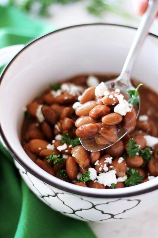A spoonful of pinto beans topped with feta cheese and parsley, bowl of pinto beans on a white table with a green napkin in the background.