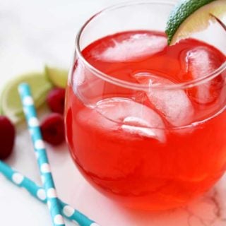 Red Singapore Sling cocktail in a glass with 4 ices cubes sitting on a white marble table, 2 blue straws, limes and raspberries.