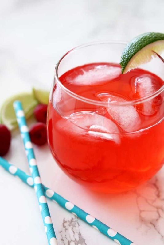 Red Singapore Sling cocktail in a clear glass with 4 ices cubes sitting on a white marble table, 2 blue straws, limes and raspberries.