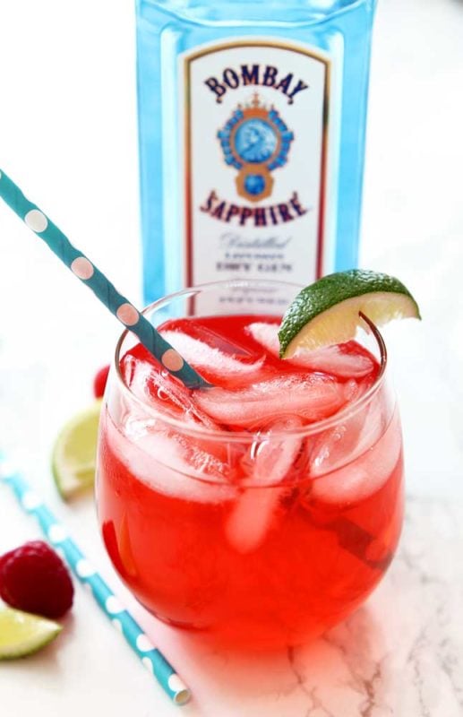 Red Singapore Sling cocktail in a glass with blue straw and lime, sitting on a white table with a blue bottle of Bombay Sapphire Gin,3 limes and a raspberry.