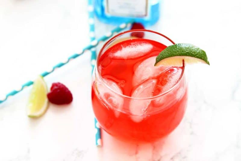 Red Singapore Sling cocktail in a glass with ice cubes sitting on a white table with a blue bottle of Bombay Sapphire Gin, 2 blue straws, limes and a raspberry.