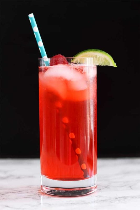 Red Singapore Sling cocktail with a blue and white straw, topped with a raspberry and lime wedge, sitting on a marble table.