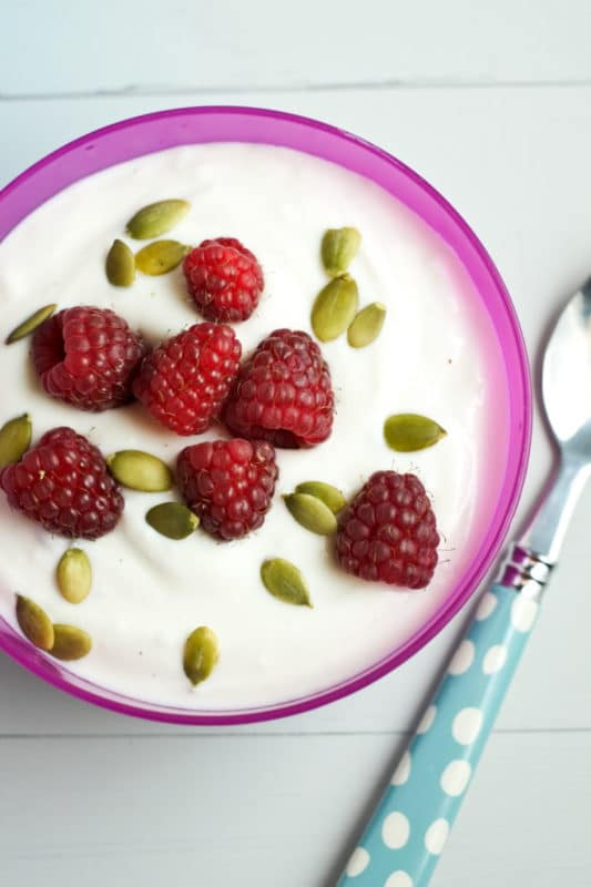 Purple bowl sitting on a white table containing plain Greek Yogurt, topped with fresh raspberries and pumpkins seeds, a blue and white polka dot spoon.