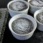Four white ramekins that contain chocolate lava cakes on a black table; topped with powdered sugar.