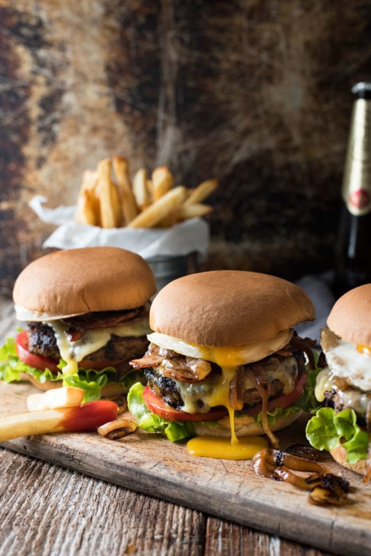 Wooden slab containing 3 cheeseburgers with melted cheese, tomatoes and lettuce, french fries in the background.